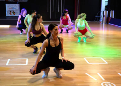 Eine Gruppe Frauen hockt beim Fitnesstraining in der Tanzschule Ritmo Latino in Ulm. Sie tragen leichte Kleidung und Turnschuhe auf einem Holzboden.
