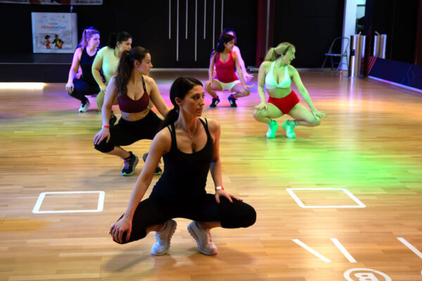 Eine Gruppe Frauen hockt beim Fitnesstraining in der Tanzschule Ritmo Latino in Ulm. Sie tragen leichte Kleidung und Turnschuhe auf einem Holzboden.