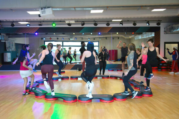 Eine Gruppe von Männern und Frauen in Sportkleidung kreist um eine Fitnessmatte in der Tanzschule Ritmo Latino in Ulm. Im Hintergrund reflektiert ein großer Spiegel sein Bild.