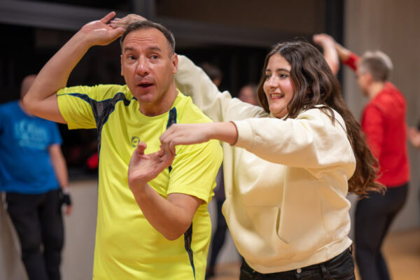 Paar Tänzer tanzen Salsa in Ulm. Sie dreht sich hinter ihm und nimmt seine Hände. Sie tragen farbenfrohe Kleidung. Im Hintergrund sind weitere Schüler der Tanzschule Ritmo Latino@ zu sehen.